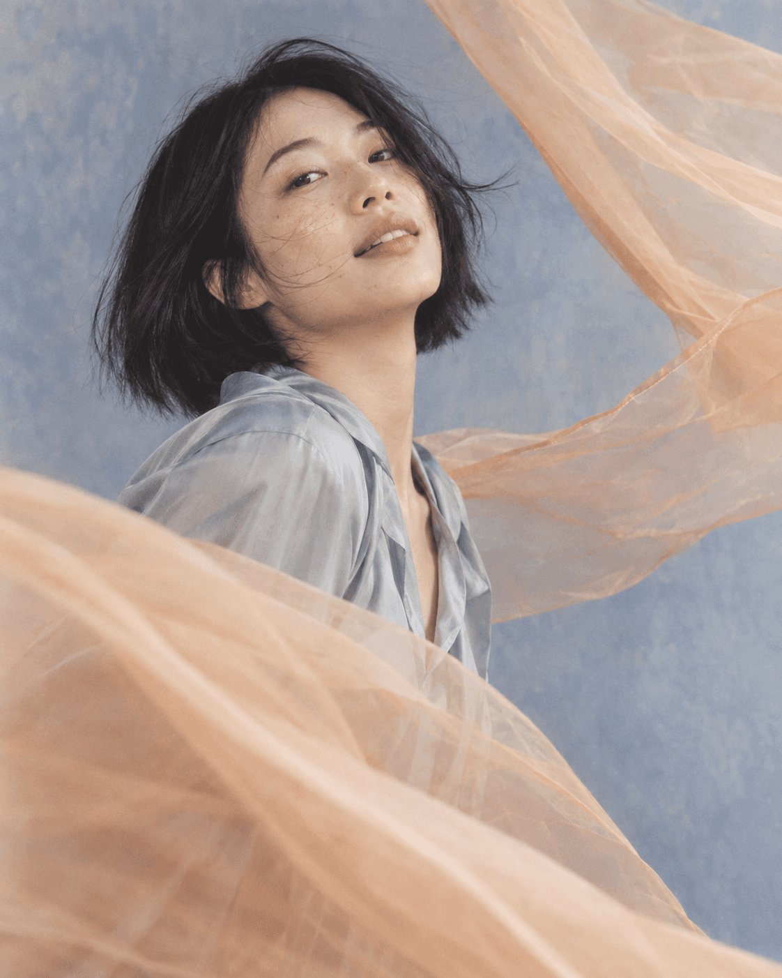 浅蓝背景中的柔和肖像，风吹黑色短发与飘动的杏色欧根纱。